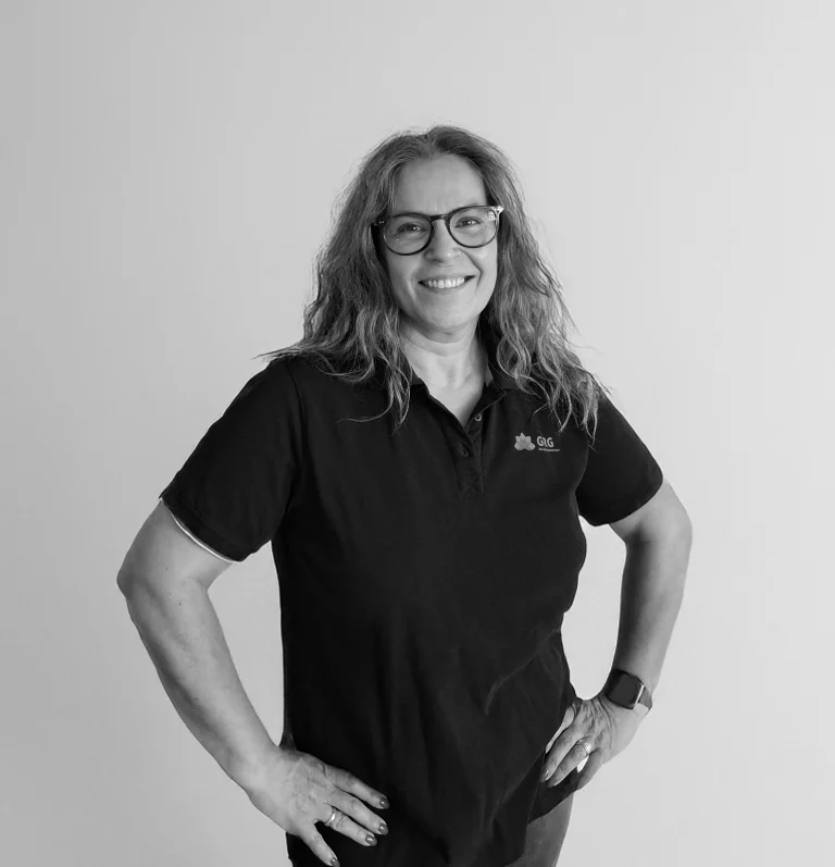 Frau mit langen blonden Haaren in einem schwarzen Poloshirt trägt eine Brille und steht vor einer neutralen Wand. Das Bild ist schwarz weiß.
