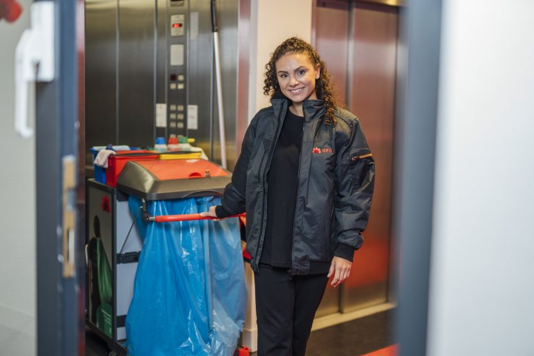 Lächelnde GRG-Mitarbeiterin in schwarzem Dienstshirt und Jacke steht an einem Reinigungswagen und steigt aus dem Auszug aus.